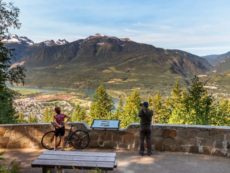 Two bikers stand on a large platform that gives a panorama view of the city of Revelstoke from Mount Revelstoke National Park