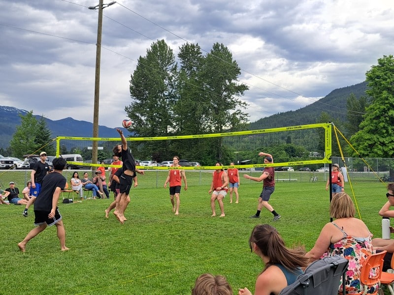 Two teams of volleyball players in a grass field with audiences on either side of the game. Part of the Devyn Gale Memorial Volleyball Tournament