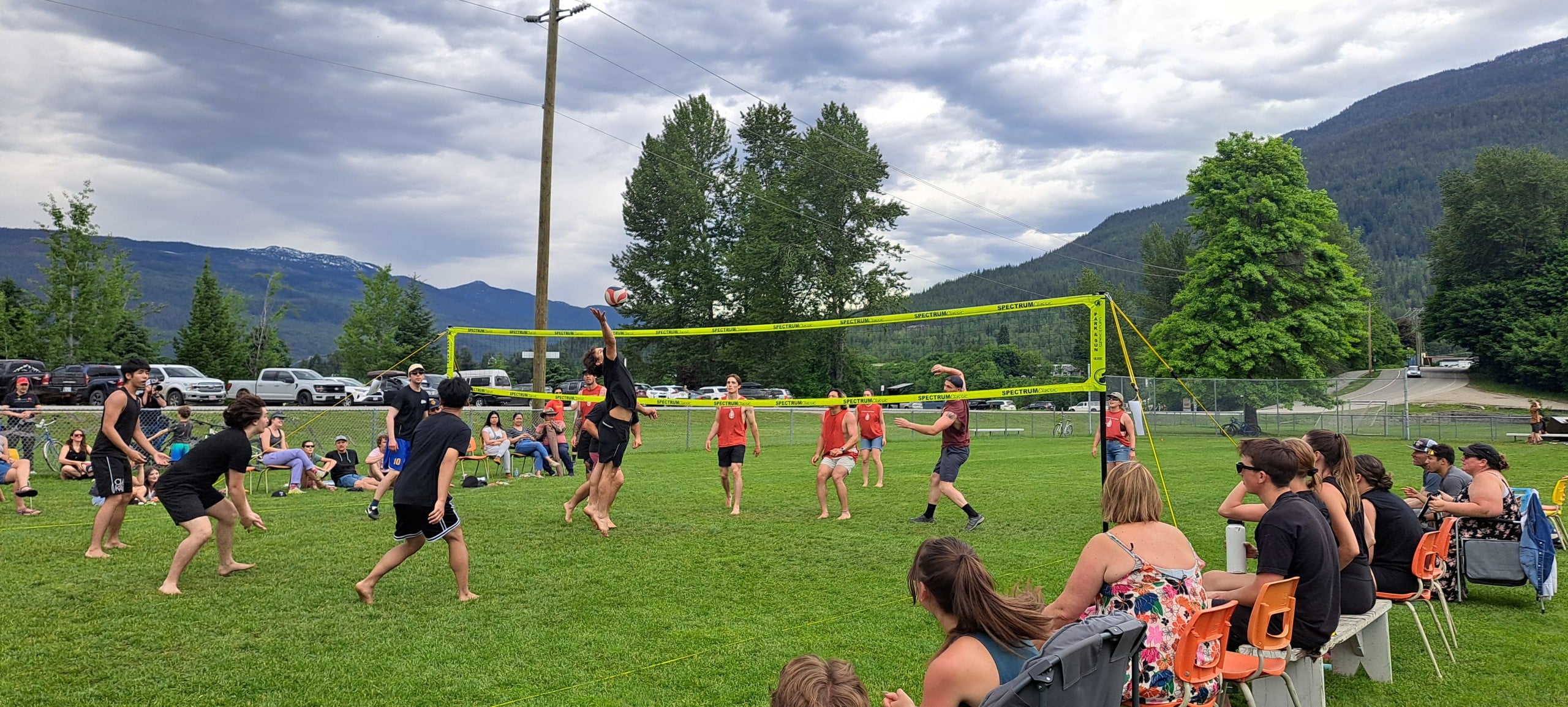 Two teams of volleyball players in a grass field with audiences on either side of the game. Part of the Devyn Gale Memorial Volleyball Tournament