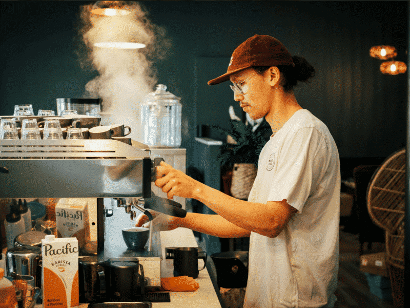 Reigan Drummond Front of House Manager for Flourish Bakery, stands at a large espresso machine and steams milk in preparation for a customer order.