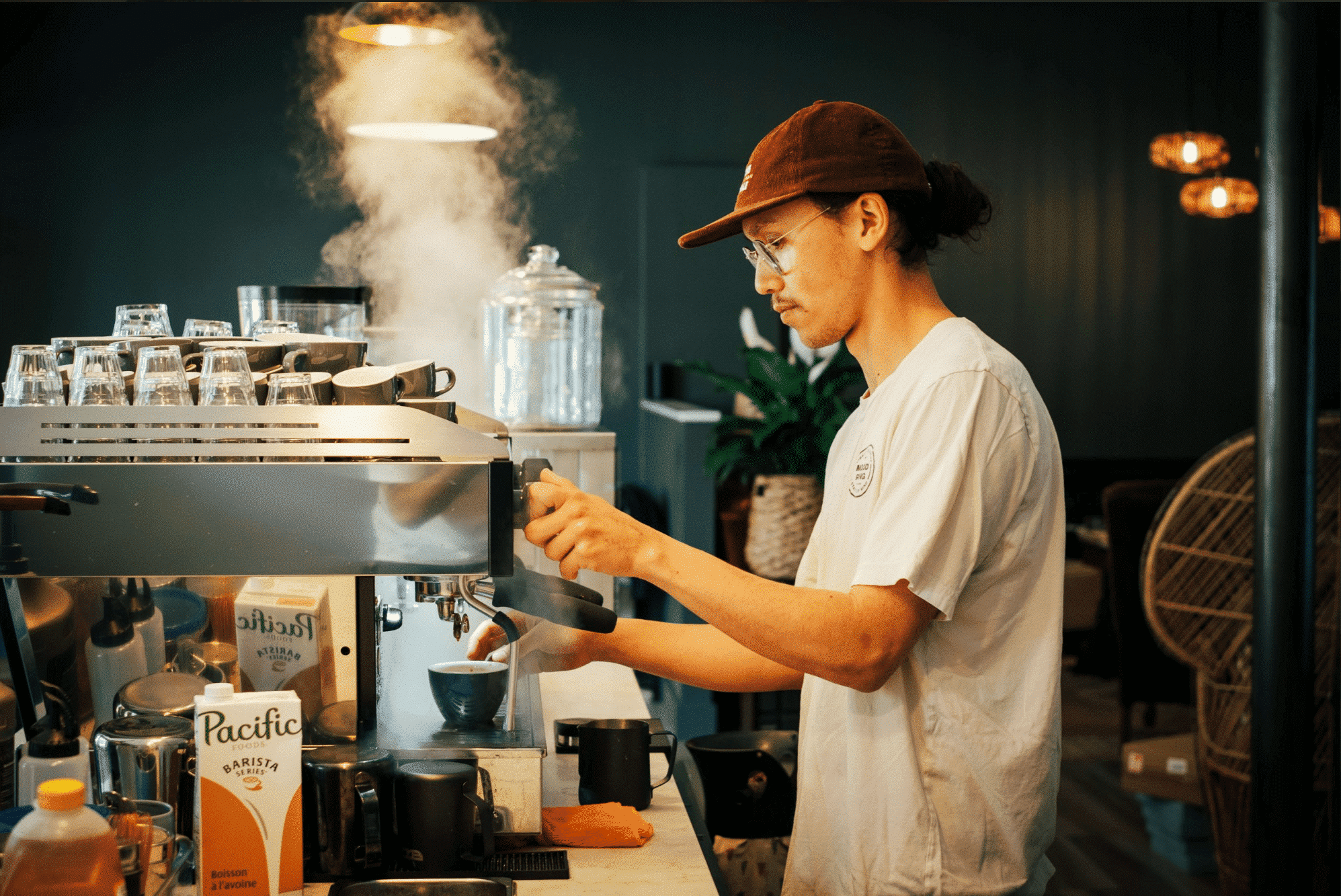 Reigan Drummond Front of House Manager for Flourish Bakery, stands at a large espresso machine and steams milk in preparation for a customer order.