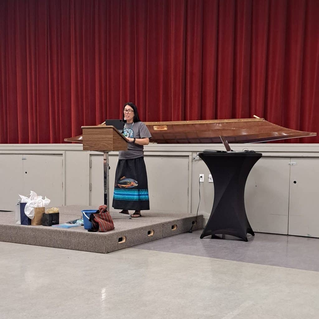 Cindy Marchand, Sinixt woman stands at a booth with a sturgeon kayak behind her as she addresses a crowd.