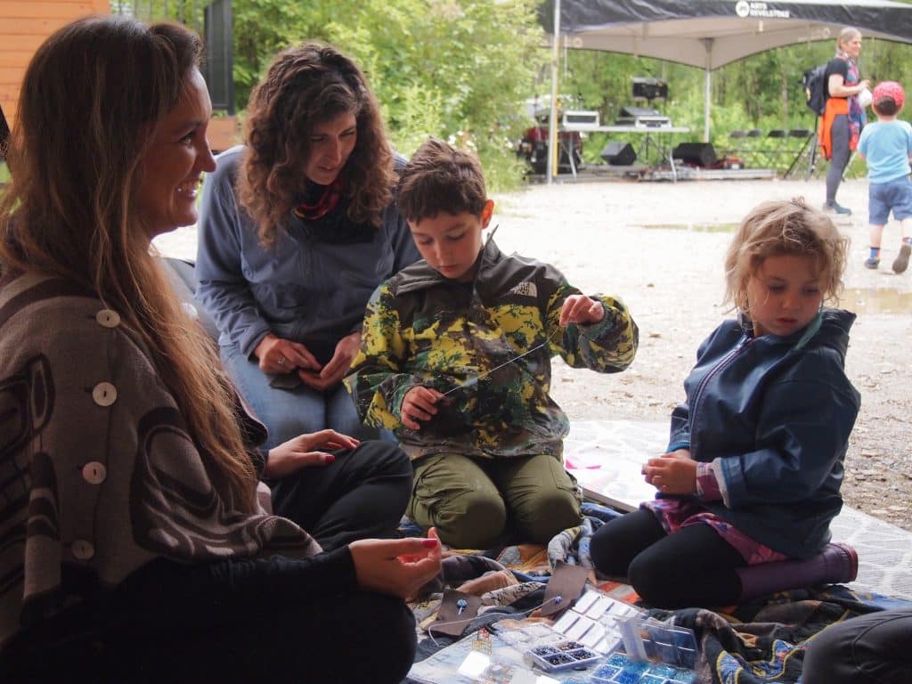 Two adults and two children sit on a carpet outside, all around a collection of beading supplies.