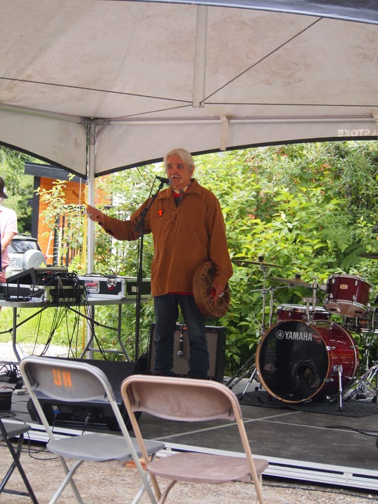 Elder Randy Williams standing on a stage surrounded by sound equipment, holding a hand drum as he addresses a crowd. He is wearing a brown leather jacket with an Orange Shirt pin.

Part of Revelstoke's National Indigenous History Month celebrations.
