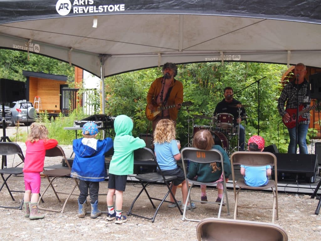 Francis Baptiste stands on a stage, holding a guitar and singing to a crowd of children all sitting and standing in the front row of chairs. Part of Revelstoke's National Indigenous History Month celebrations.