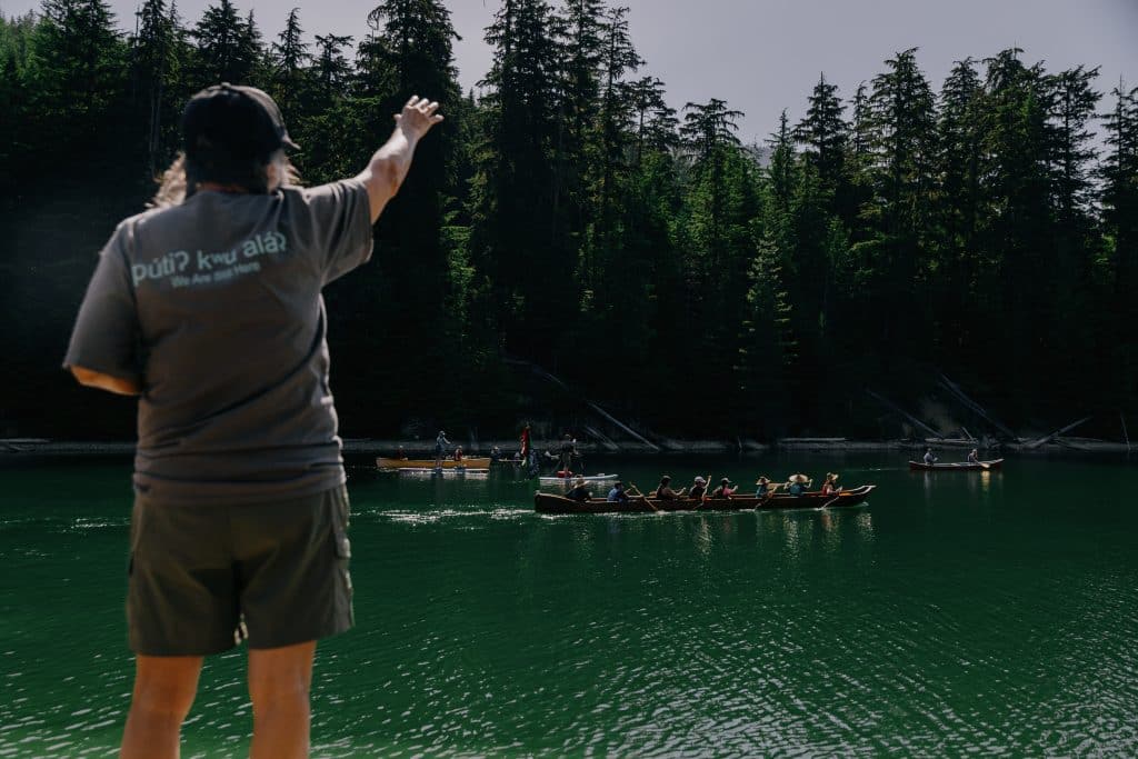 Sinixt Elder Shelly Boyd standing on a rocky outcropping, hand raised as she looks over a collection of canoes, kayaks and paddle boards floating on the Columbia River where it pools into Lake Revelstoke dam reservoir.

Part of Revelstoke's National Indigenous History Month celebrations.