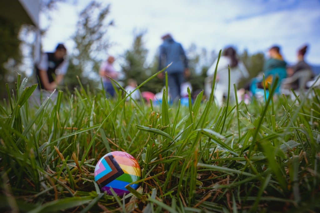 Mountain Pride 2025 promotion image of a pride pin in the grass with a crowd of participants out of focus in the background