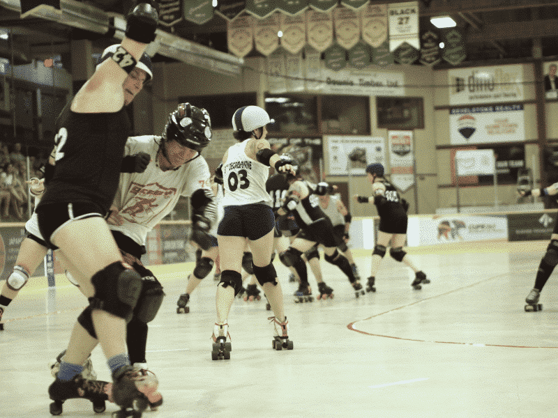 Over six roller derby players in the middle of a bout. Two players at the front of the pack are staking backwards. Two of the players are from the Revelstoke Derailers