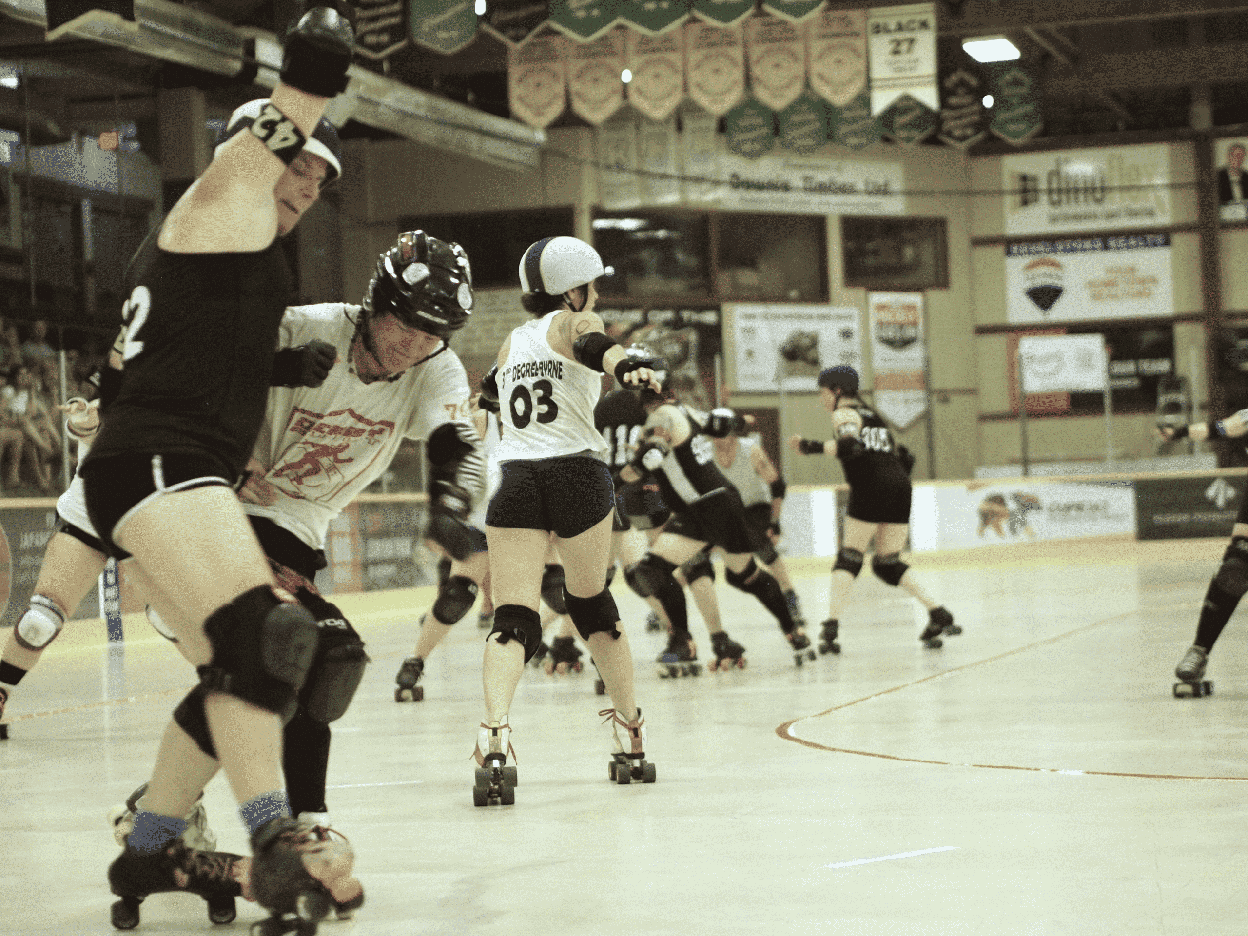Over six roller derby players in the middle of a bout. Two players at the front of the pack are staking backwards. Two of the players are from the Revelstoke Derailers