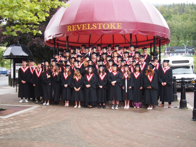 Revelstoke Class of 2025 in graduation gowns all lining up in front of the Revelstoke Grizzly band shell