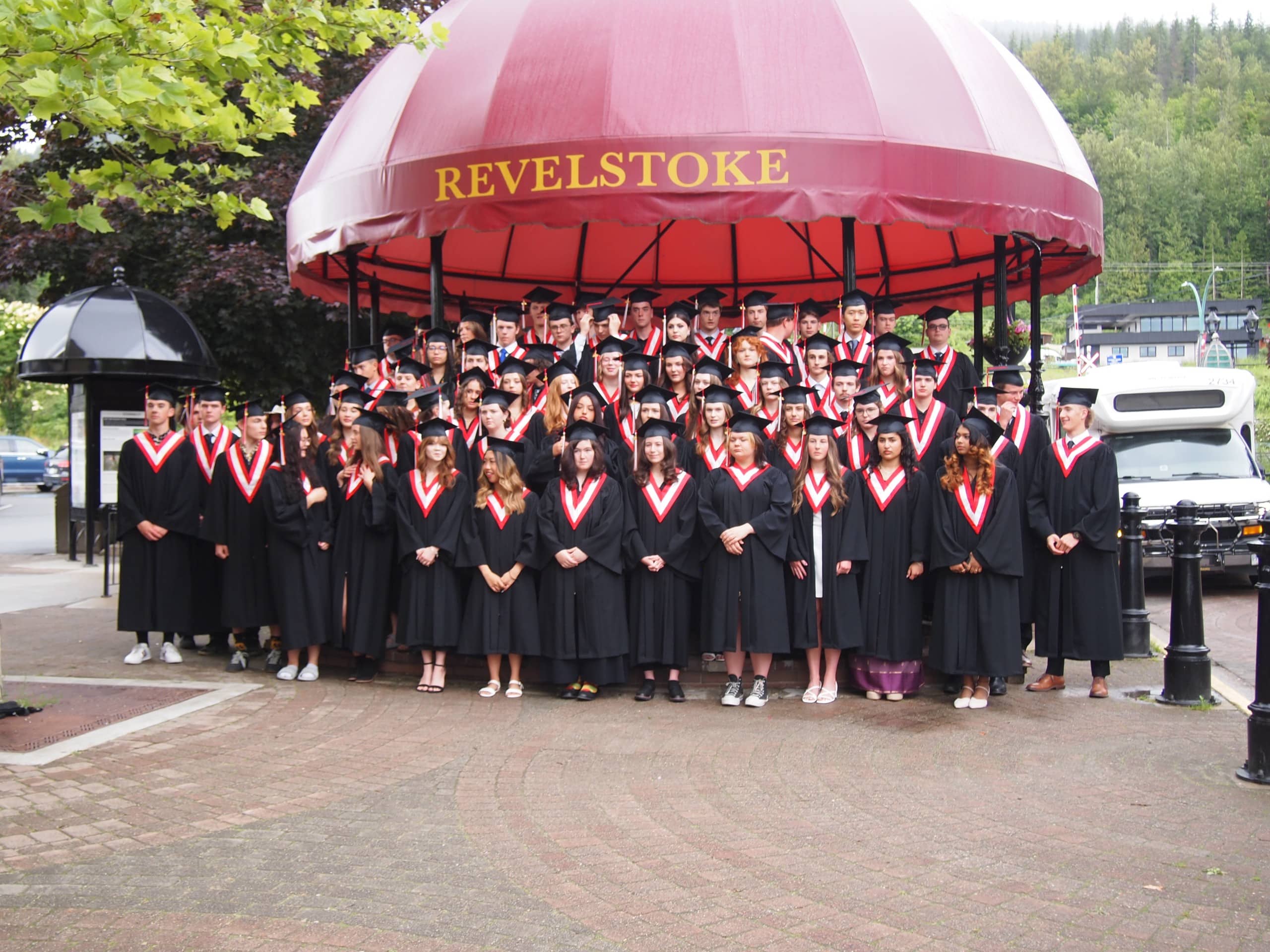 Revelstoke Class of 2025 in graduation gowns all lining up in front of the Revelstoke Grizzly band shell