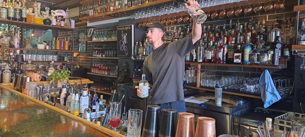 Monashee Spirits bartender Jesse Moore standing behind the bar lined with bottles as he holds up a quarter filled glass bottle, showing ingredients to customers.