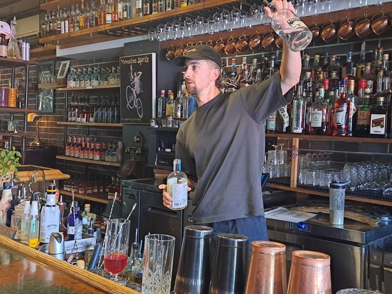 Monashee Spirits bartender Jesse Moore standing behind the bar lined with bottles as he holds up a quarter filled glass bottle, showing ingredients to customers.
