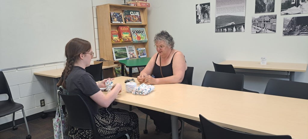 Two individuals at a table, one of them working on a crochet piece as they both chat.