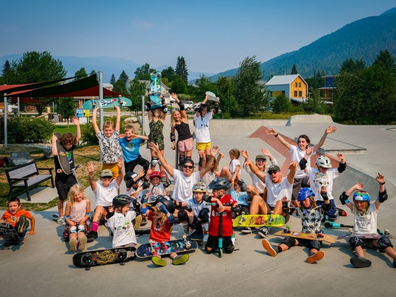 A collection of Stoked Skate Camp participants standing by the playground at Kovach skate park, looking at the camera and smiling or cheering. Some are holding up their skateboards.