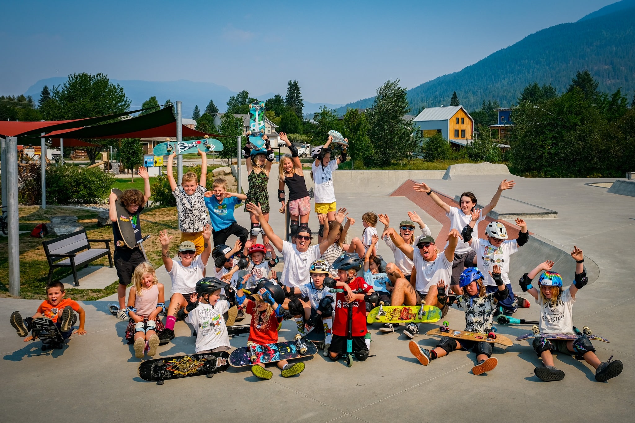 A collection of Stoked Skate Camp participants standing by the playground at Kovach skate park, looking at the camera and smiling or cheering. Some are holding up their skateboards.
