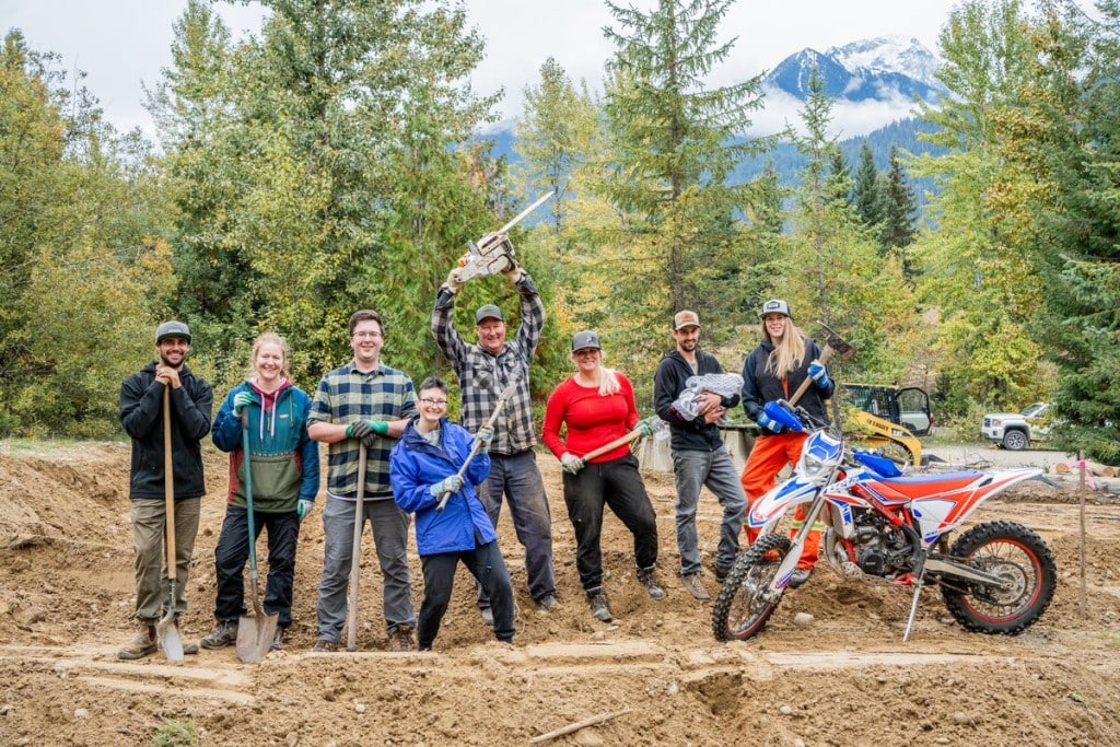 A group of Thanksgiving Back volunteers stand side by side on a large dirt trail, some holding shovels as they look at the camera and smile.