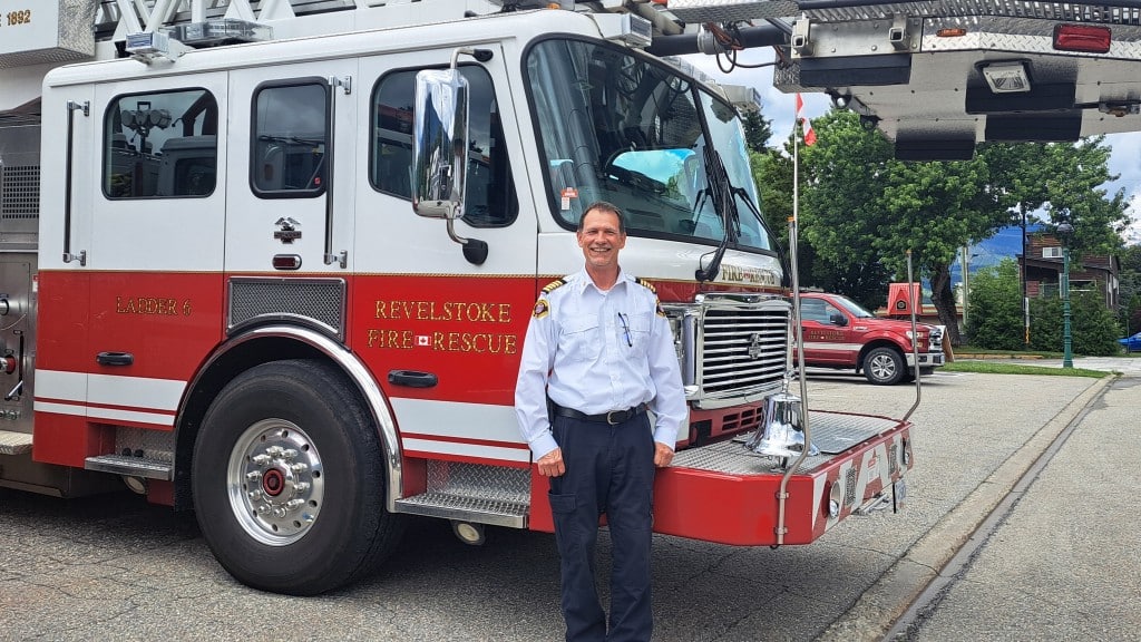Steven DeRousie, Revelstoke Fire Rescue Services fire chief stands in front of a Revelstoke fire rescue engine wearing a white uniform shirt and black pants. He is looking at the camera and smiling,