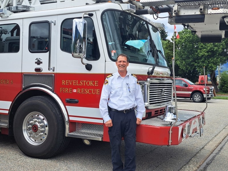 Steven DeRousie, Revelstoke Fire Rescue Services fire chief stands in front of a Revelstoke fire rescue engine wearing a white uniform shirt and black pants. He is looking at the camera and smiling,