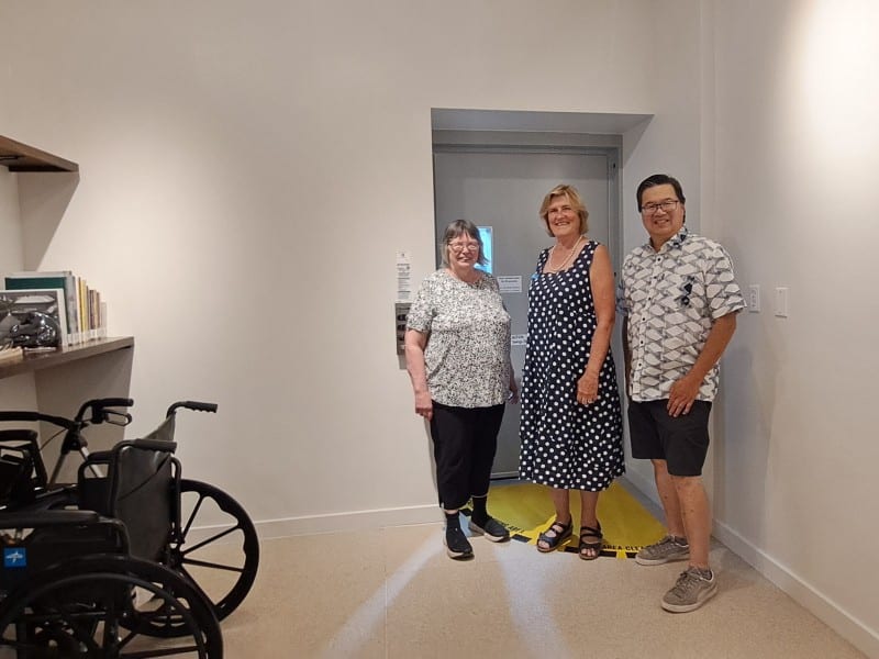 Three people stand in front of the metal doors of an access lift in a brightly light room at the back of the Revelstoke Museum and Archives. The door is the entrance to the new accessibility lift.
