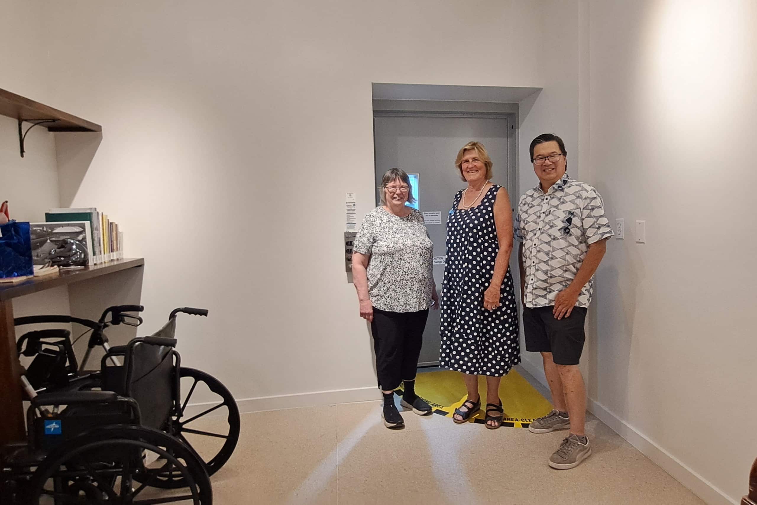 Three people stand in front of the metal doors of an access lift in a brightly light room at the back of the Revelstoke Museum and Archives. The door is the entrance to the new accessibility lift.