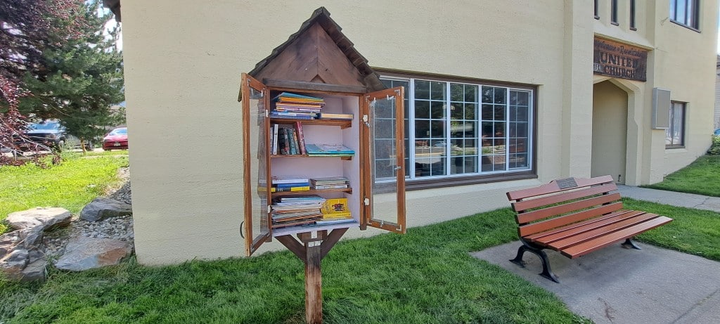 Revelstoke united church little free library, a large wooden box in the style of a house with two doors swinging open to show four shelves of books. The doors have plexiglass in the middle. 