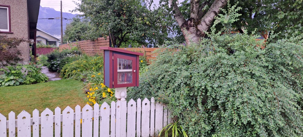 A small red, wooden little free library behind a white picket fence