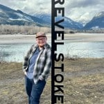 Revelstoke Mountaineer's community journalist Lys Morton, a white man with a shaved head and a small brown beard stands leaning against a metal Revelstoke sign with the Columbia river and a mountain range behind him. He is smiling at the camera.
