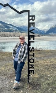 Revelstoke Mountaineer's community journalist Lys Morton, a white man with a shaved head and a small brown beard stands leaning against a metal Revelstoke sign with the Columbia river and a mountain range behind him. He is smiling at the camera.