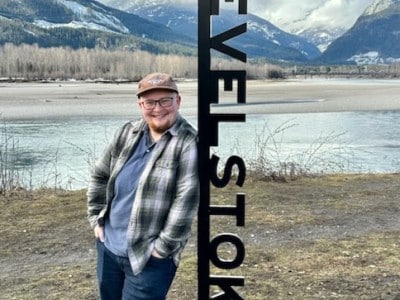 Revelstoke Mountaineer's community journalist Lys Morton, a white man with a shaved head and a small brown beard stands leaning against a metal Revelstoke sign with the Columbia river and a mountain range behind him. He is smiling at the camera.