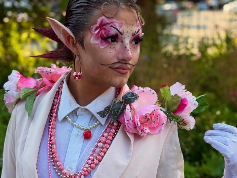 Jackie Daytona in a cream white suit with pink and striped tiger lily flowers on his collar and lapels. Long, prosthetic fairy ears, makeup highlightng the eyebrows and strong, hot pink eyeshadow on the face accentuating the brow lines.
