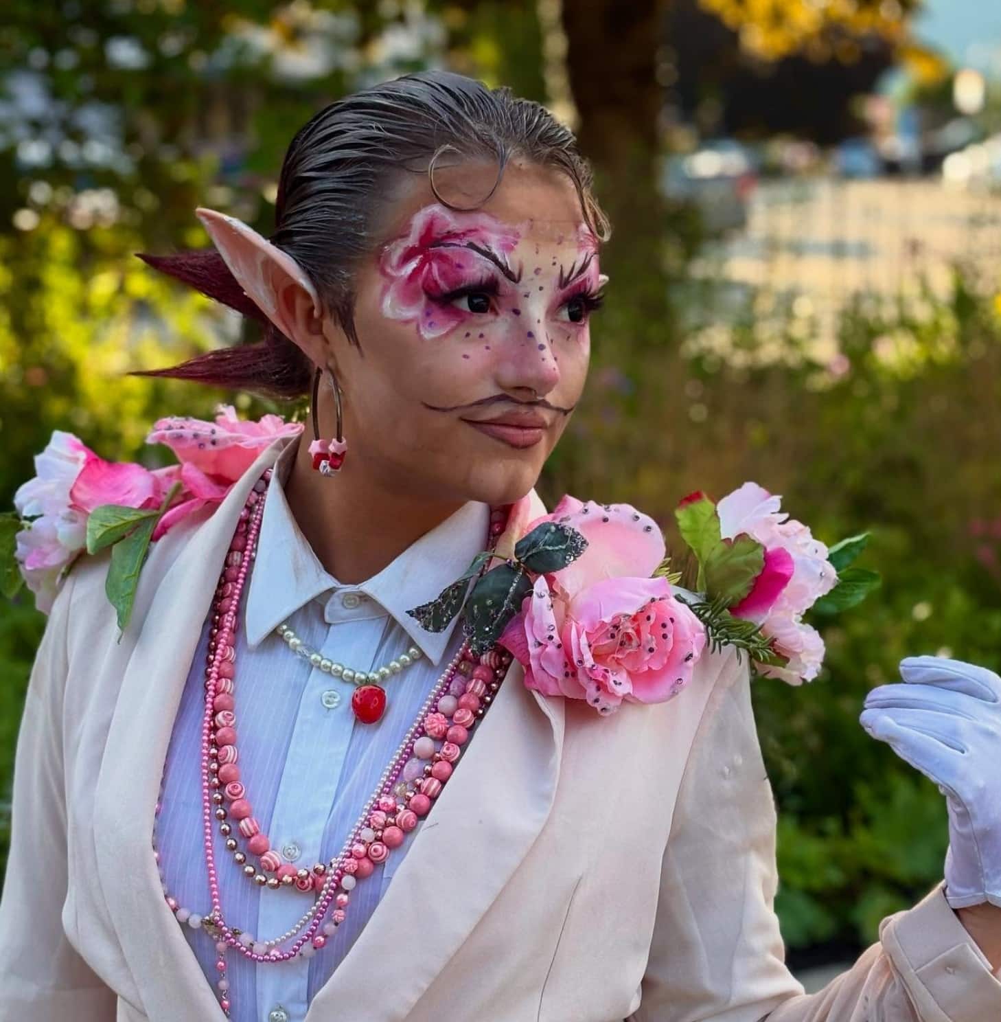Jackie Daytona in a cream white suit with pink and striped tiger lily flowers on his collar and lapels. Long, prosthetic fairy ears, makeup highlightng the eyebrows and strong, hot pink eyeshadow on the face accentuating the brow lines.