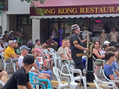 Saxophone player for the artist Wesli walking through the crowd at Revy.Live Outside as he plays. People are sitting in white plastic chairs, many smiling as they all watch the performance.
