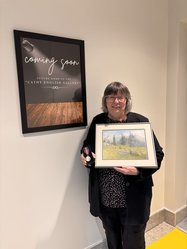 Cathy English standing in front of the Cathy English Gallery signage holding a medal and a painting.