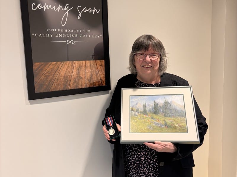 Cathy English standing in front of the Cathy English Gallery signage holding a medal and a painting.