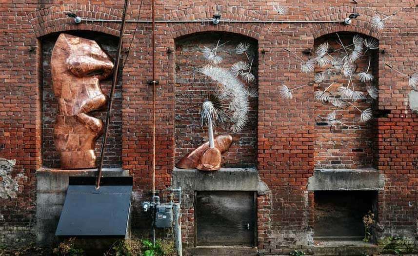 Revelstoke Art Alleries piece A Simple Joy, a metalwork face blowing the seeds off a dandelion head as the seeds are attached to different sections of a brick wall.