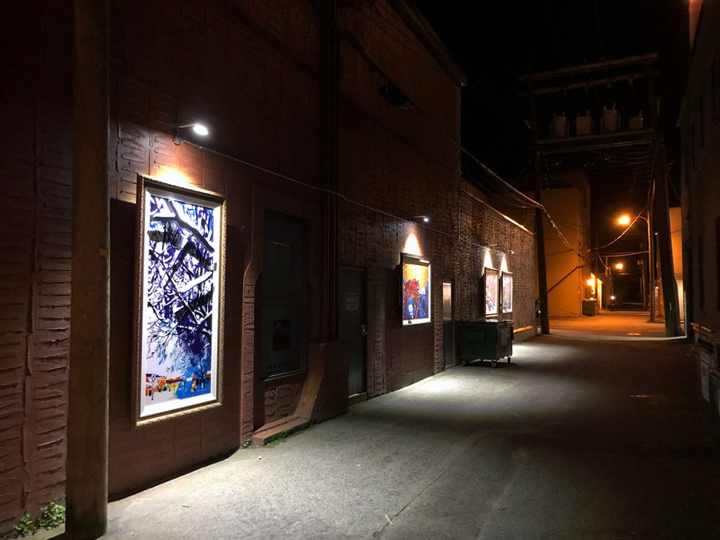 Revelstoke Art Alleries pieces Contemporary Landscape, four different metal works of different landscapes around Revelstoke. All of them on a brick wall, lit up with LED overhead light.