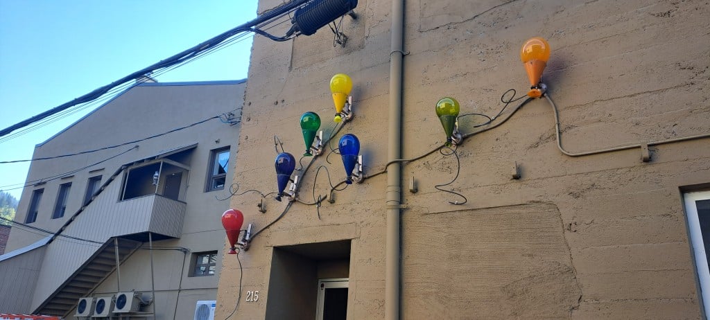 A collection of glass balloons in different colours connected to metal "string" as they all float up the side of the building.
