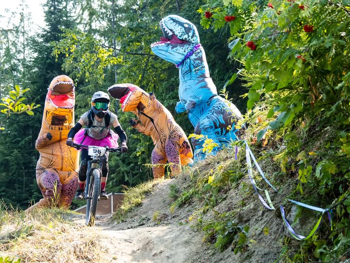 Revelstoke Women’s Enduro racer coming down the path on a mountain bike while three audience members dressed in inflatable dinosaur costumes sheer from the sideline.