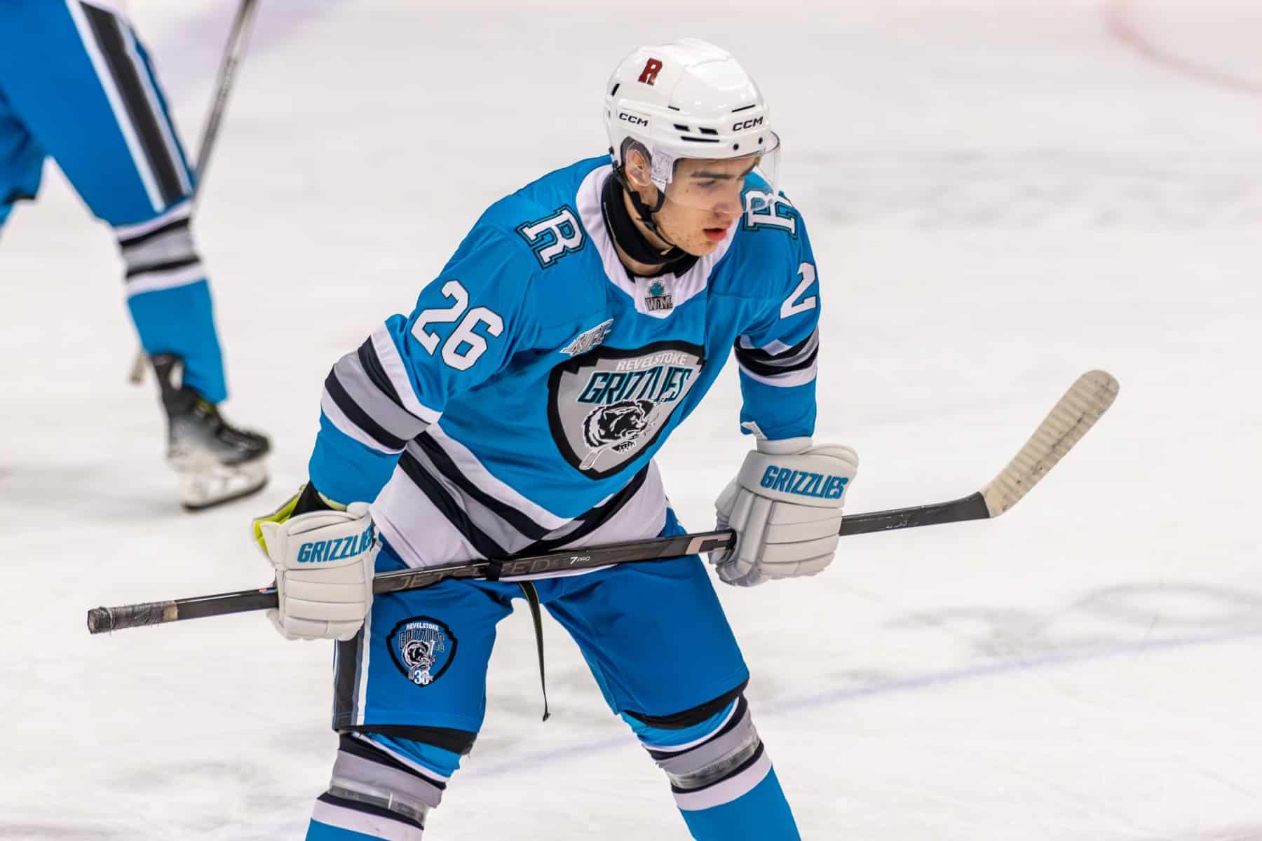 Connor Shymoniak, Revelstoke Grizzlies player in the blue Revelstoke Grizzlies jersey. He is on the ice, staring towards the other end, hands on knees and stick braced.