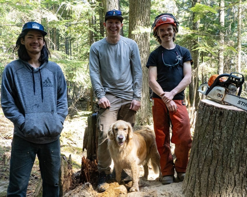 Revelstoke Cycling Association 2025 trail crew, three men standing in a forest with tree cutting gear around them and a bike leaning against a tree stump.