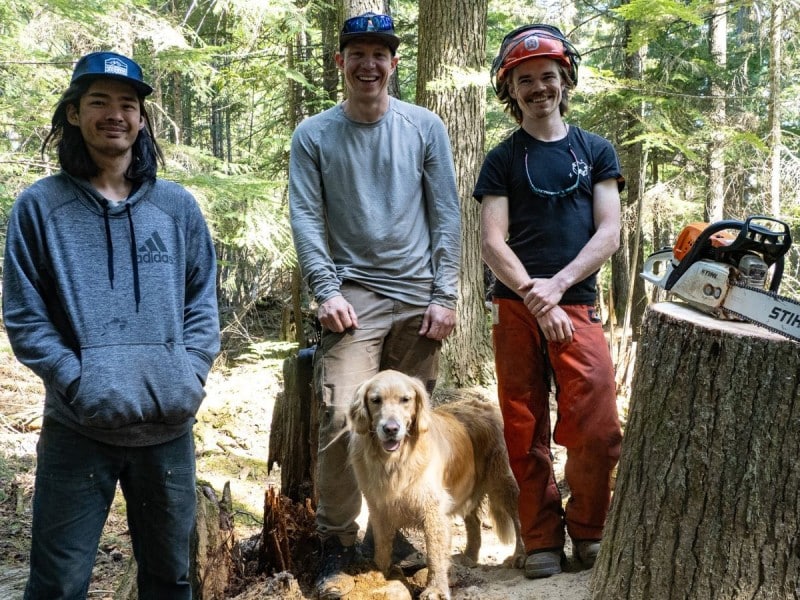 Revelstoke Cycling Association 2025 trail crew, three men standing in a forest with tree cutting gear around them and a bike leaning against a tree stump.