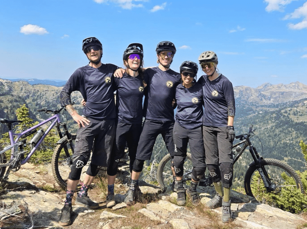 Jackalope Bikes ride team, five peoiple all dressed in biking gear and helmets, stand in a group on a ridge, back to the drop and two bikes. All are looking at the camera and smiling.