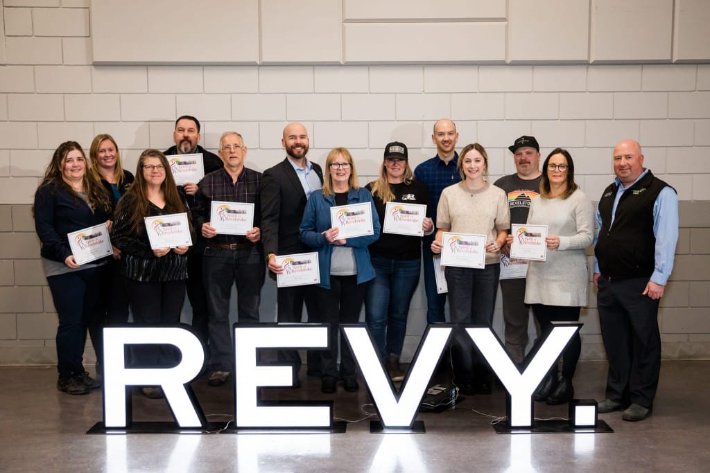 12 Spirit of Revelstoke award winners standing behind a lit up REVY. sign, all displaying their award certificates.