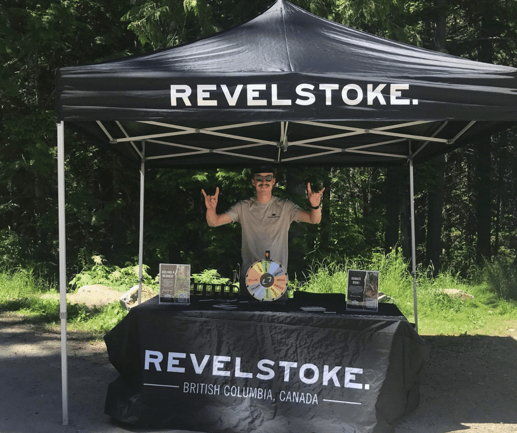 Tourism Revelstoke Stoke Team member standing behind a Tourism Revelstoke branded table, holding up Downtown Dollars