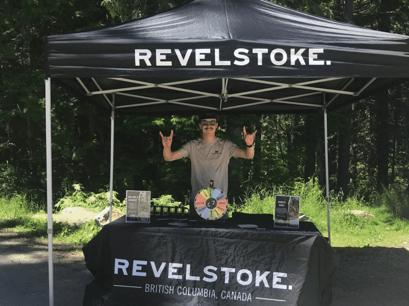Tourism Revelstoke Stoke Team member standing behind a Tourism Revelstoke branded table, holding up Downtown Dollars