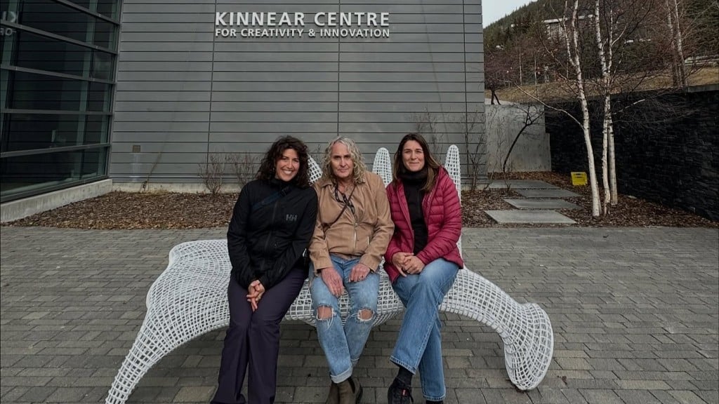 Three women sitting on a white wire bench outside the Banff art centre