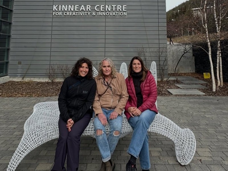 Three women sitting on a white wire bench outside the Banff art centre
