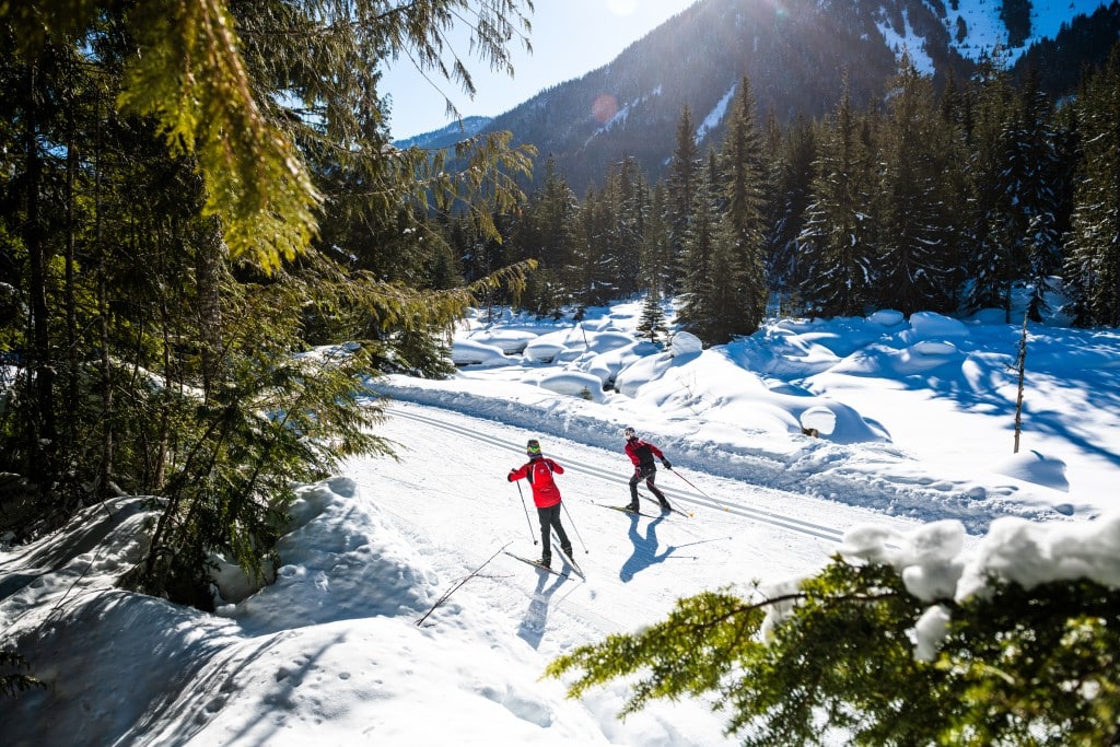 Revelstoke Nordic Ski Club members on a groomed trail part of the Mount Macpherson route. The sun is shining and the cross country skiers are crossing a break between trees.