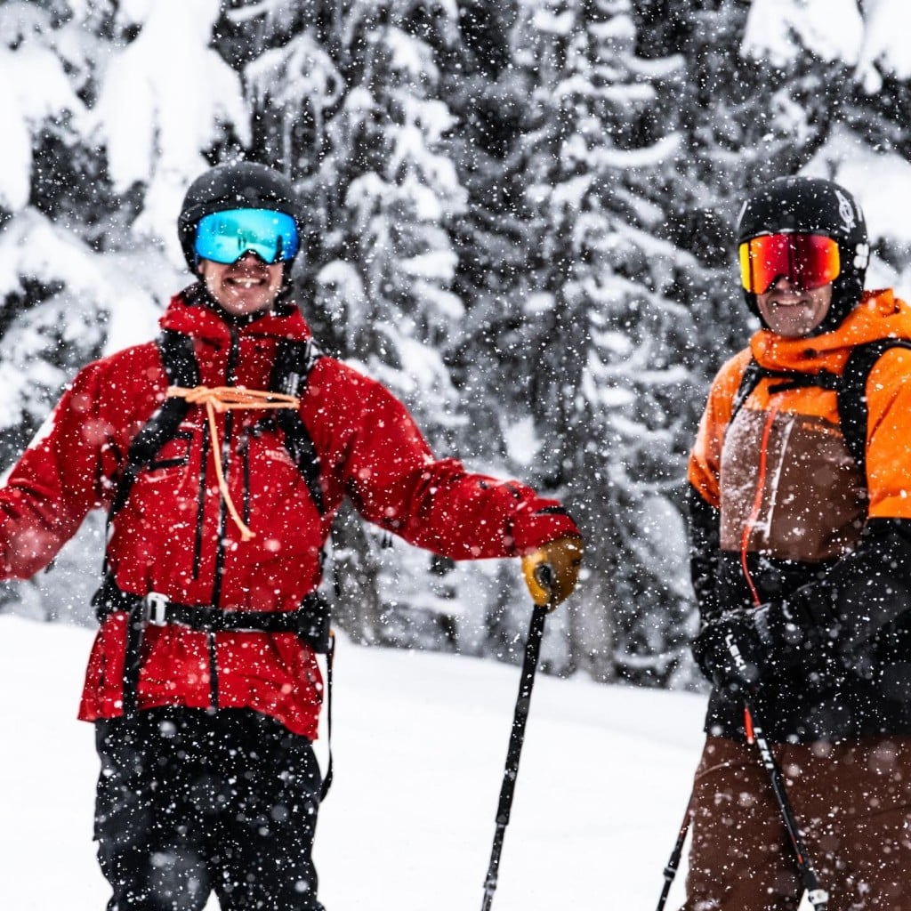 Revelstoke Mountain Resort opening day 2025 guests, two of them in ski gear and holding ski poles as they look at the camera and smile. Snow is noticeably falling.